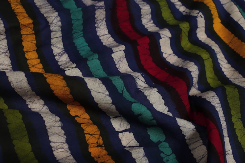 May include: Close-up of a blue fabric with white, black, orange, green, and red stripes. The fabric has a batik pattern with a textured, crinkled look.
