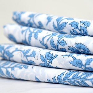 Op de afbeelding: Een stapel gevouwen witte stof met een herhalend blauw bloemenpatroon. Het ontwerp toont gestileerde bladeren en wijnranken. De stof is strak gevouwen, wat de ingewikkelde details van het patroon en het contrast tussen blauw en wit laat zien.