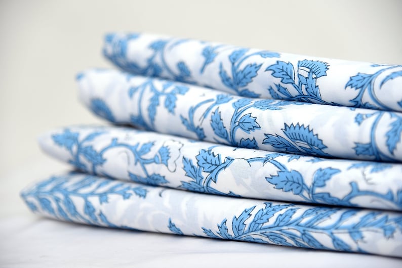Op de afbeelding: Een stapel gevouwen witte stof met een herhalend blauw bloemenpatroon. Het ontwerp toont gestileerde bladeren en wijnranken. De stof is strak gevouwen, wat de ingewikkelde details van het patroon en het contrast tussen blauw en wit laat zien.