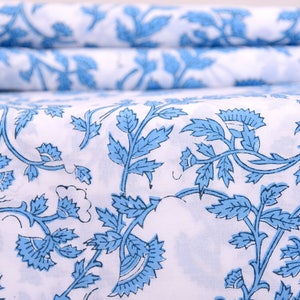 Op de afbeelding: Witte stof met een herhalend patroon van blauwe bloemenranken en bladeren. De stof is gevouwen, met het patroon zichtbaar op de bovenste laag en de gevouwen randen. Het ontwerp is een klassieke, elegante stijl, geschikt voor diverse woondecoratieprojecten.