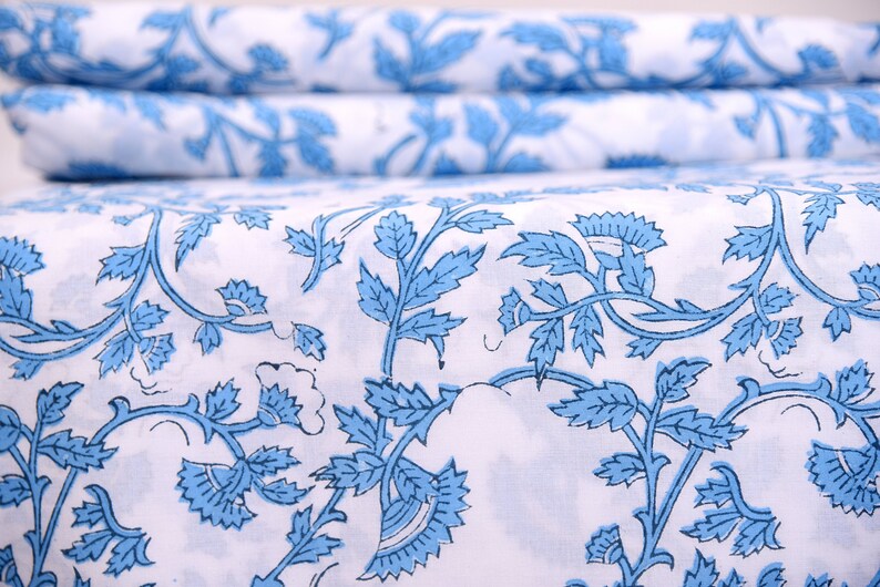 Op de afbeelding: Witte stof met een herhalend patroon van blauwe bloemenranken en bladeren. De stof is gevouwen, met het patroon zichtbaar op de bovenste laag en de gevouwen randen. Het ontwerp is een klassieke, elegante stijl, geschikt voor diverse woondecoratieprojecten.
