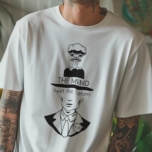 May include: White t-shirt with a black graphic design featuring a head with a mushroom cloud above it and the text "THE MIND split the atom". The shirt is worn by a person with tattoos.