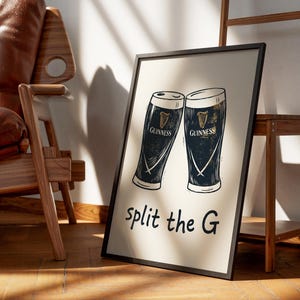 May include: A framed print featuring two stylized Guinness pint glasses. The glasses are black and white with gold accents and the Guinness logo. The phrase "split the G" is written in black script below the glasses.