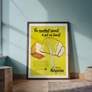 May include: A framed vintage margarine advertisement. The poster has a yellow background with the text "The smoothest spread to put on bread." Slices of bread are shown with margarine being spread on them. The poster is in a brown wooden frame.