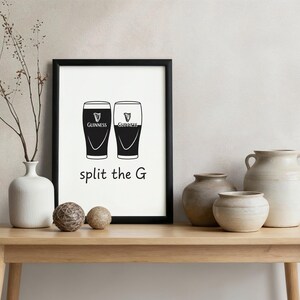 May include: A framed print with two black Guinness pint glasses, the text "split the G" below. The print is in a black frame, displayed on a wooden table with decorative vases and spheres.