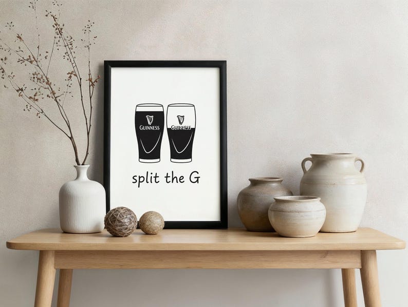 May include: A framed print with two black Guinness pint glasses, the text "split the G" below. The print is in a black frame, displayed on a wooden table with decorative vases and spheres.