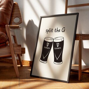 May include: Black framed print featuring two stylized Guinness pint glasses with the text "split the G". The glasses are black and white, with the Guinness logo. The print is leaning against a wall, near a wooden chair and a wooden shelf.