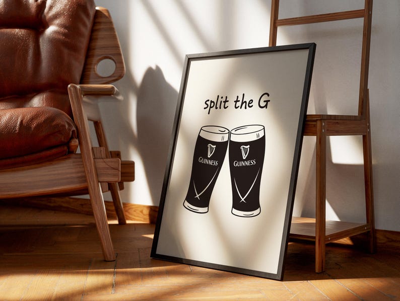 May include: Black framed print featuring two stylized Guinness pint glasses with the text "split the G". The glasses are black and white, with the Guinness logo. The print is leaning against a wall, near a wooden chair and a wooden shelf.