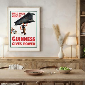 May include: A framed vintage Guinness advertisement. The poster features a worker carrying a steel beam with the text "Build Your Muscles" and "Guinness Gives Power" in red. A seated figure is also depicted.