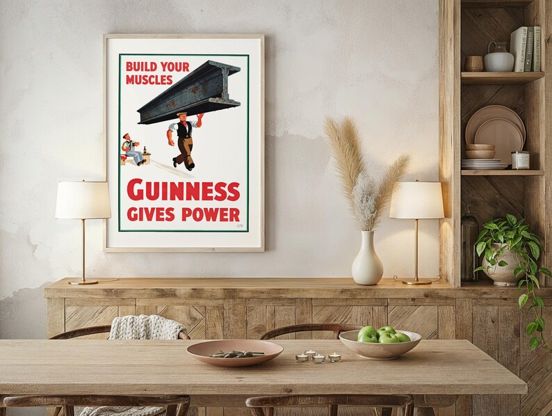 May include: A framed vintage Guinness advertisement. The poster features a worker carrying a steel beam with the text "Build Your Muscles" and "Guinness Gives Power" in red. A seated figure is also depicted.