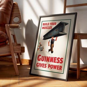 May include: Framed vintage Guinness advertisement. The poster features a man carrying a steel beam with the text "Build Your Muscles" and "Guinness Gives Power" in red. A second man is seated at a table. The frame is black.