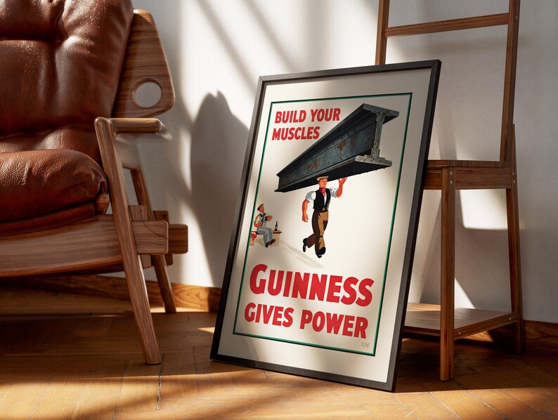 May include: Framed vintage Guinness advertisement. The poster features a man carrying a steel beam with the text "Build Your Muscles" and "Guinness Gives Power" in red. A second man is seated at a table. The frame is black.