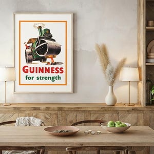 May include: Framed vintage Guinness advertisement poster. The poster shows a green steamroller with the text "Guinness for strength" in red and green. The poster is displayed on a wall above a wooden table with a lamp.