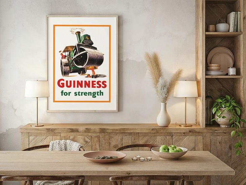 May include: Framed vintage Guinness advertisement poster. The poster shows a green steamroller with the text "Guinness for strength" in red and green. The poster is displayed on a wall above a wooden table with a lamp.
