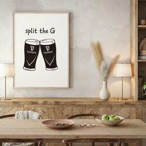 May include: A framed print featuring two black Guinness pint glasses with the text "split the G" above them. The print is displayed on a wooden cabinet in a dining room setting, with a table, lamps, and decorative items.
