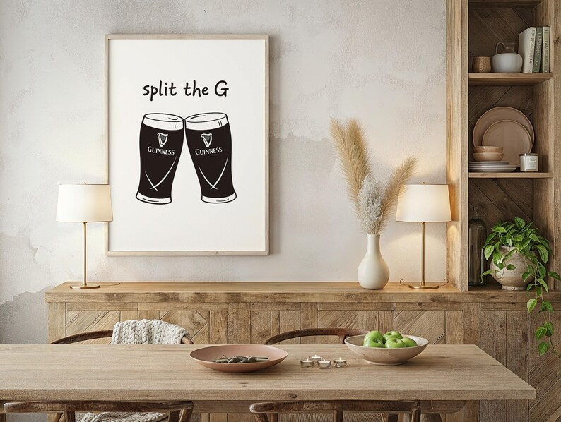 May include: A framed print featuring two black Guinness pint glasses with the text "split the G" above them. The print is displayed on a wooden cabinet in a dining room setting, with a table, lamps, and decorative items.