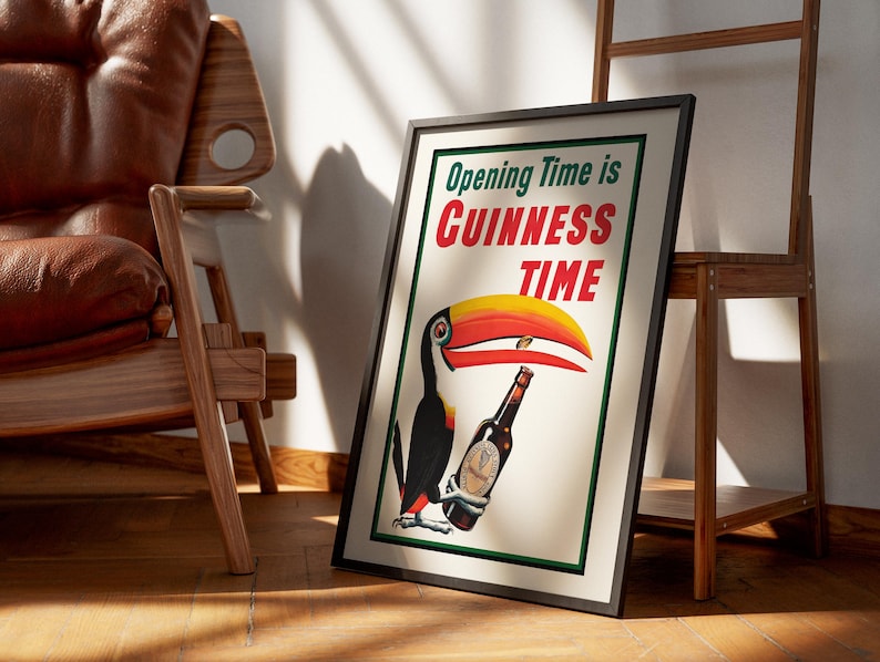 Puede incluir: P&oacute;ster enmarcado con borde negro que presenta un tuc&aacute;n sosteniendo una botella de Guinness. El p&oacute;ster dice "Opening Time is Guinness Time" en texto verde y rojo. La imagen est&aacute; apoyada contra una escalera de madera y una silla de cuero marr&oacute;n.