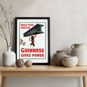May include: A framed vintage Guinness advertisement. The poster features a man carrying a steel beam with the text "Build Your Muscles" and "Guinness Gives Power" in red and white. The poster is displayed on a wooden table with decorative vases.