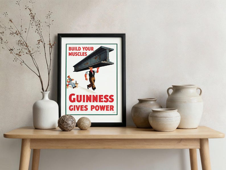 May include: A framed vintage Guinness advertisement. The poster features a man carrying a steel beam with the text "Build Your Muscles" and "Guinness Gives Power" in red and white. The poster is displayed on a wooden table with decorative vases.
