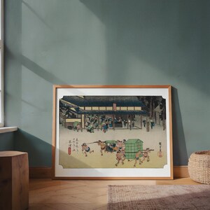 May include: Framed Japanese woodblock print depicting a bustling outdoor scene with figures carrying objects. The print features a building with a dark roof and numerous people. The artwork is in a wooden frame and rests on a wooden floor.