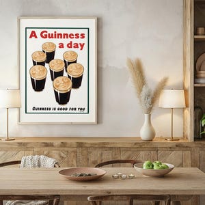 May include: Framed vintage-style print featuring six pints of Guinness with creamy foam. The text reads "A Guinness a day" in red and "Guinness is good for you" in black. The print is displayed on a wooden table with lamps and decorative items.