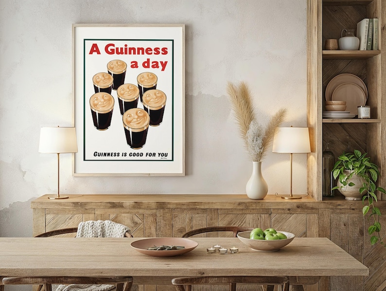 May include: Framed vintage-style print featuring six pints of Guinness with creamy foam. The text reads "A Guinness a day" in red and "Guinness is good for you" in black. The print is displayed on a wooden table with lamps and decorative items.