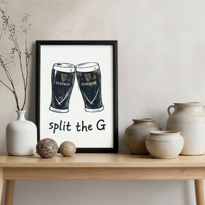 May include: Black framed print featuring two Guinness pint glasses with the text "split the G". The print is displayed on a light wood table with decorative vases and spheres.