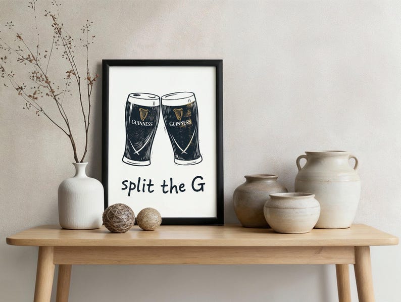 May include: Black framed print featuring two Guinness pint glasses with the text "split the G". The print is displayed on a light wood table with decorative vases and spheres.