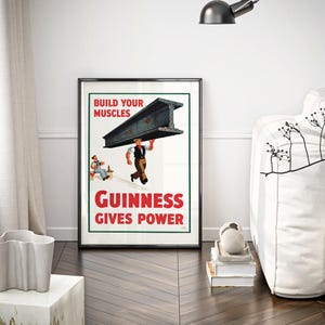 May include: A framed vintage Guinness advertisement poster. The poster features a man carrying a steel beam with the text "BUILD YOUR MUSCLES" and "GUINNESS GIVES POWER" in red. The poster is displayed in a room with a white sofa and a decorative vase.