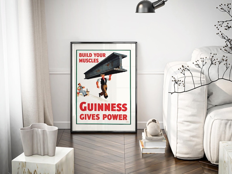 May include: A framed vintage Guinness advertisement poster. The poster features a man carrying a steel beam with the text "BUILD YOUR MUSCLES" and "GUINNESS GIVES POWER" in red. The poster is displayed in a room with a white sofa and a decorative vase.