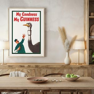 May include: Framed vintage advertisement poster with the text "My Goodness My Guinness" in red and white. The artwork features a cartoon of a man and an ostrich. The poster is displayed above a wooden table.