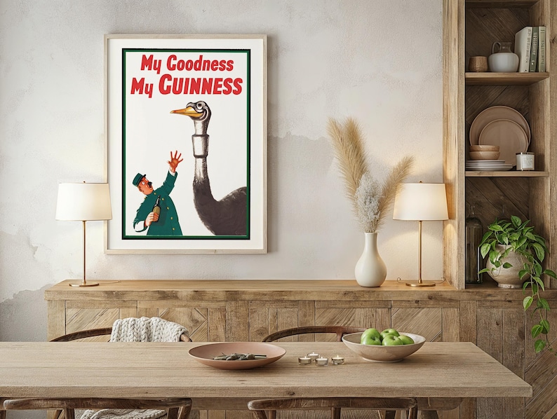 May include: Framed vintage advertisement poster with the text "My Goodness My Guinness" in red and white. The artwork features a cartoon of a man and an ostrich. The poster is displayed above a wooden table.
