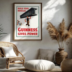 May include: A framed vintage Guinness advertisement poster. The poster features a worker carrying a steel beam with the text "BUILD YOUR MUSCLES" and "GUINNESS GIVES POWER" in red. The poster is displayed in a room with a wicker chair and decorative vase.