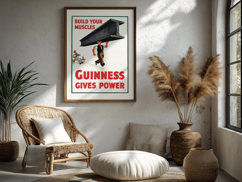May include: A framed vintage Guinness advertisement poster. The poster features a worker carrying a steel beam with the text "BUILD YOUR MUSCLES" and "GUINNESS GIVES POWER" in red. The poster is displayed in a room with a wicker chair and decorative vase.