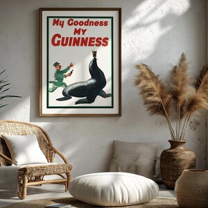 May include: A framed vintage Guinness advertisement featuring a seal balancing a pint of Guinness. The poster has the text "My Goodness My Guinness" in red and white. The artwork is set in a room with a wicker chair, plants, and decorative vases.