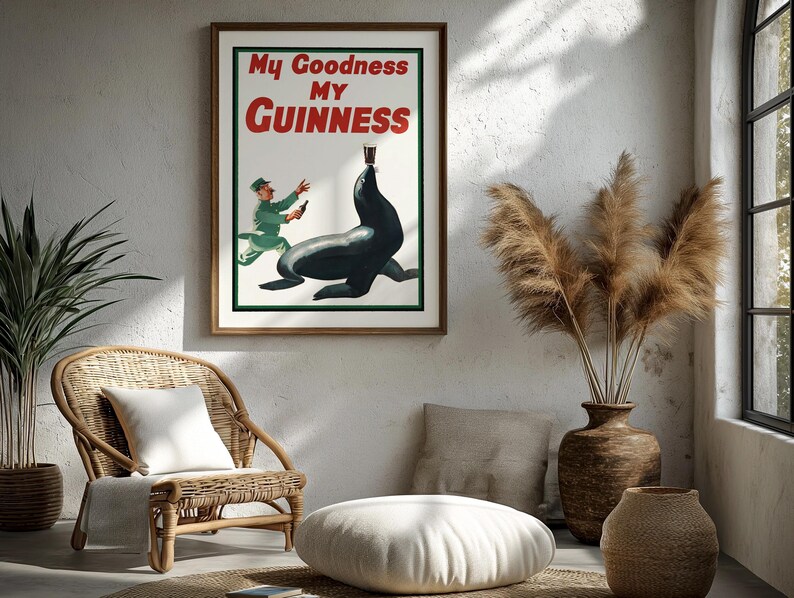 May include: A framed vintage Guinness advertisement featuring a seal balancing a pint of Guinness. The poster has the text "My Goodness My Guinness" in red and white. The artwork is set in a room with a wicker chair, plants, and decorative vases.