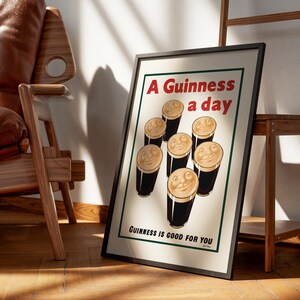 May include: Framed vintage-style poster with the text "A Guinness a day" and "Guinness is good for you." The poster features cartoon pints of Guinness with smiling faces. The poster is displayed on a wooden floor, leaning against a wall.