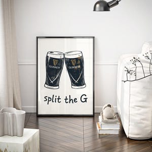 May include: Black framed art print featuring two Guinness pint glasses with the Guinness logo. The text "split the G" is below the glasses. The print is displayed in a room with a white sofa and a decorative plant.