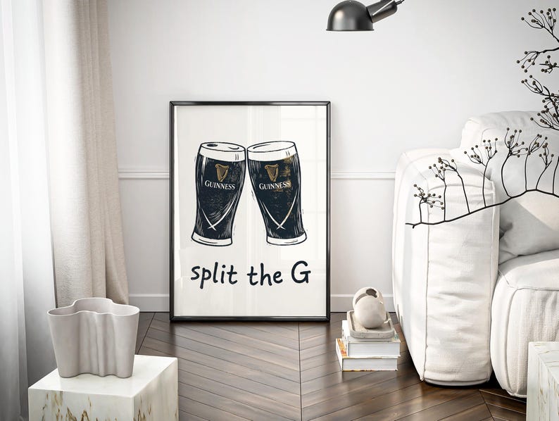 May include: Black framed art print featuring two Guinness pint glasses with the Guinness logo. The text "split the G" is below the glasses. The print is displayed in a room with a white sofa and a decorative plant.