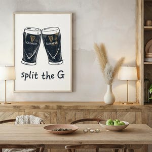 May include: A framed print featuring two Guinness pint glasses in a black and white illustration. The text "split the G" is below the glasses. The print is displayed on a wooden sideboard in a dining room setting.