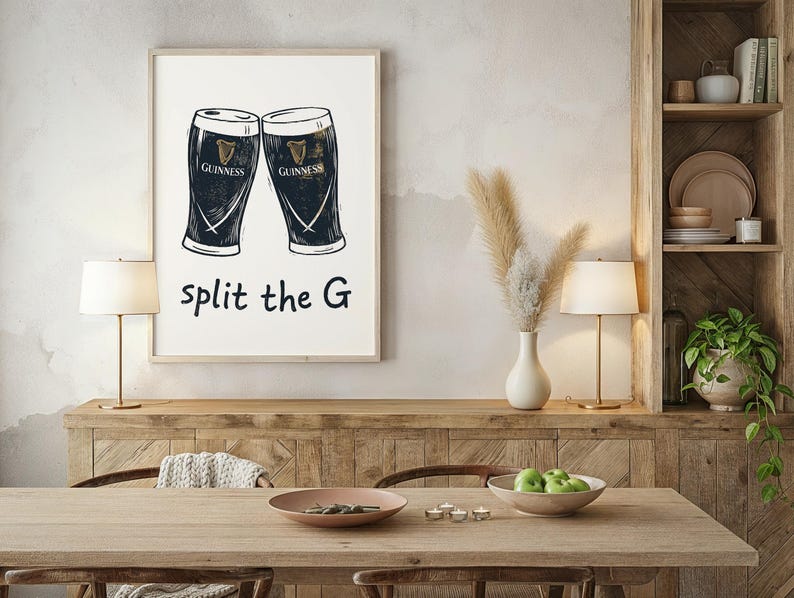 May include: A framed print featuring two Guinness pint glasses in a black and white illustration. The text "split the G" is below the glasses. The print is displayed on a wooden sideboard in a dining room setting.