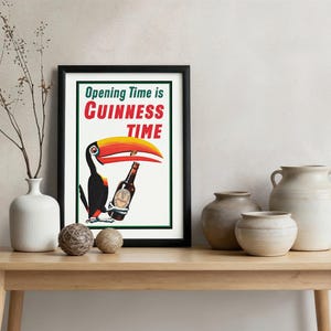 Puede incluir: Un cuadro enmarcado con borde negro que muestra un tuc&aacute;n sosteniendo una botella de Guinness. El texto "Opening Time is Guinness Time" est&aacute; en verde, rojo y blanco. El cuadro est&aacute; sobre una mesa de madera con jarrones y esferas.