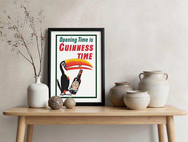Puede incluir: Un cuadro enmarcado con borde negro que muestra un tuc&aacute;n sosteniendo una botella de Guinness. El texto "Opening Time is Guinness Time" est&aacute; en verde, rojo y blanco. El cuadro est&aacute; sobre una mesa de madera con jarrones y esferas.