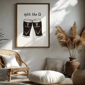 May include: A framed print featuring two Guinness pint glasses with the text "split the G". The glasses are black with white details and the Guinness logo. The print is in a wooden frame, displayed in a room with a wicker chair and decorative vase.