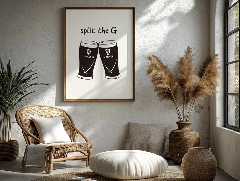 May include: A framed print featuring two Guinness pint glasses with the text "split the G". The glasses are black with white details and the Guinness logo. The print is in a wooden frame, displayed in a room with a wicker chair and decorative vase.