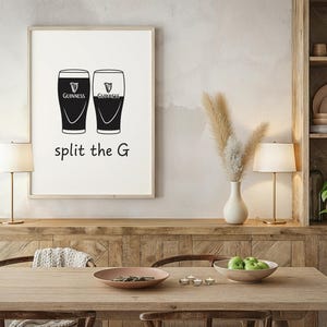 May include: A framed print featuring two stylized Guinness pint glasses, one black and one white, with the Guinness logo. Below the glasses, the text reads "split the G". The print is displayed in a room with a wooden table and a decorative shelf.