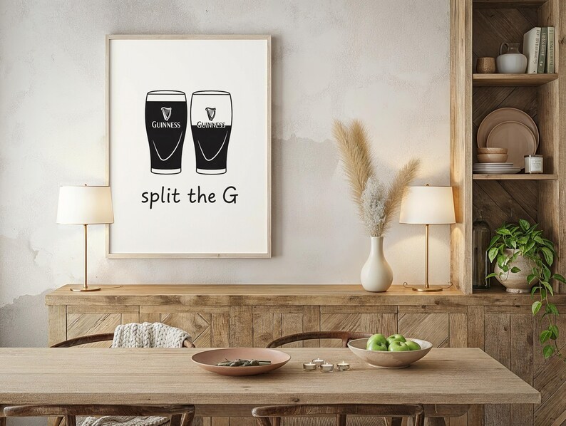 May include: A framed print featuring two stylized Guinness pint glasses, one black and one white, with the Guinness logo. Below the glasses, the text reads "split the G". The print is displayed in a room with a wooden table and a decorative shelf.