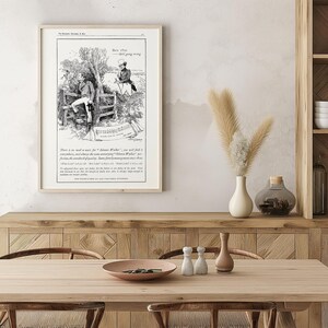 May include: Framed vintage black and white print featuring an illustration of two men in a rural setting. The print includes text and is displayed on a wooden table with decorative items. The room has a neutral color palette.