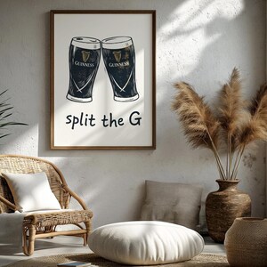 May include: A framed print featuring two Guinness pint glasses with the text "split the G". The artwork is in black and white, with the Guinness logo visible on each glass. The print is displayed in a well-lit room with a wicker chair and decorative vase.