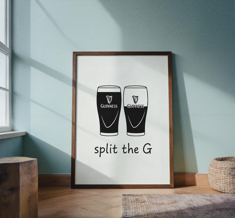 May include: A framed print featuring two Guinness pint glasses, one filled and one empty, with the text "split the G" in a black font. The Guinness logo is visible on each glass. The frame is brown.